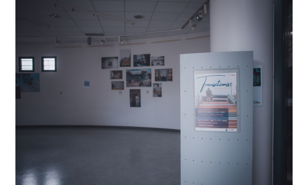 En arrière-plan, des photos accrochées à un mur. Au premier plan, un panneau avec l'affiche de l'exposition.