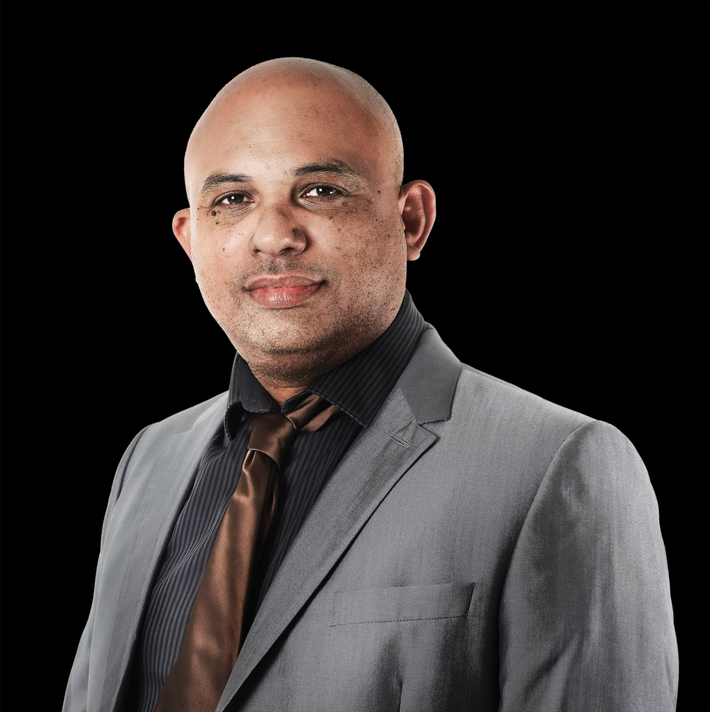 Portrait of Dr Dominique MORAU. He wears a gray suit jacket, a black shirt and a brown tie. the background of the photo is black. He stands slightly in profile and looks toward the camera