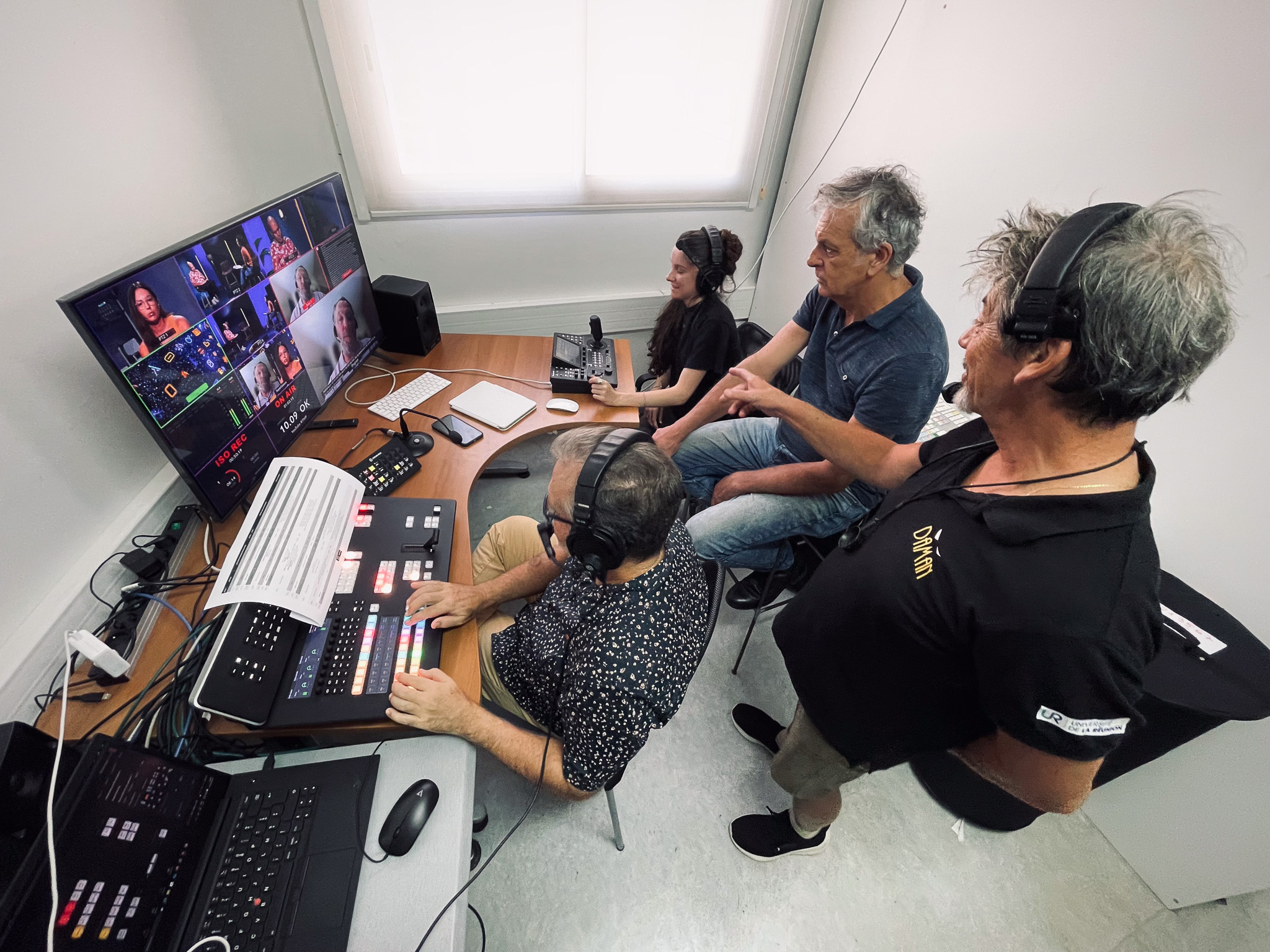 Équipe technique en régie vidéo en pleine production. Quatre personnes portent un casque audio et travaillent autour d’un grand écran de contrôle diffusant plusieurs visages en visioconférence. Une personne est aux commandes de la console de mixage vidéo, pendant que les autres échangent debout ou assis. La scène se déroule dans une salle blanche avec lumière naturelle.