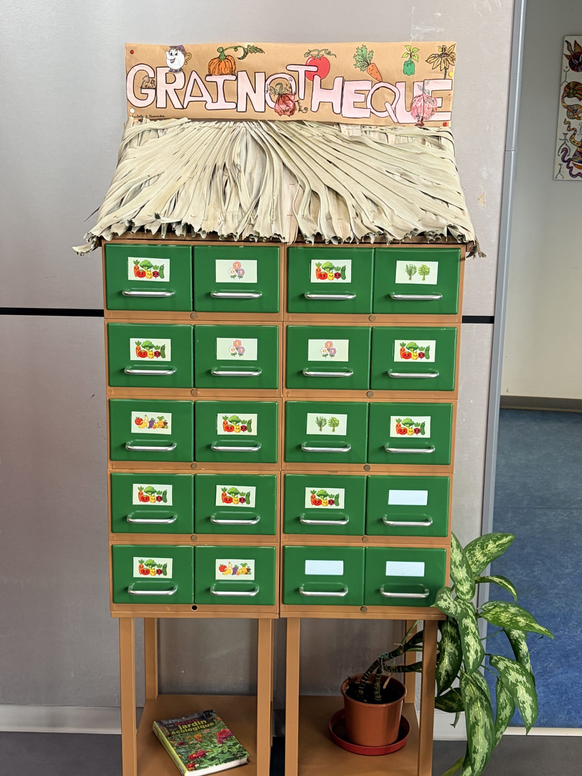 Un meuble en bois nommé « Grainothèque » composé de petits tiroirs verts illustrés de légumes et surmonté d'un toit en paille.